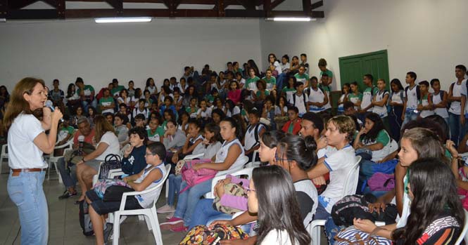 Foto visão lateral. Servidora Adriana Passos palestrando em escola. Pátio lotado de estudantes sentados em cadeiras brancas de plástico.
