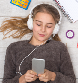 Menina deitada no chão, com livros e materiais espalhados, ouvindo áudio no celular com fones de ouvido.