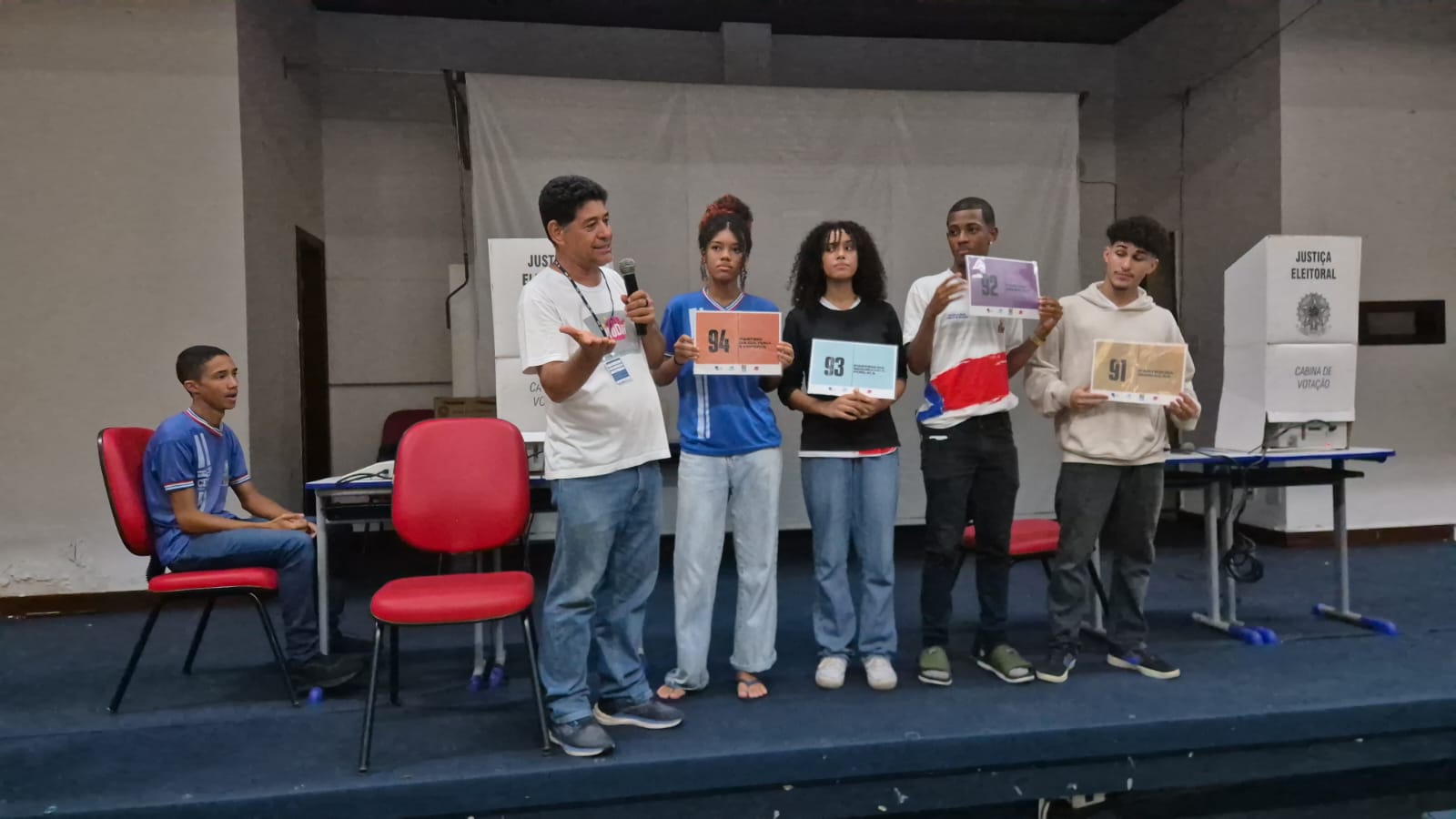 Em um palco, um pessoa fala no microfone, quatro seguram uma placa com números e outra está sentada a direita. 
