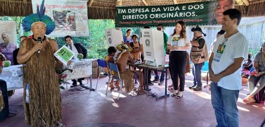 Pra todos verem: Um homem indígena segura um microfone; outro repousa as mãos; ao fundo, jovens simulam o voto em urna.