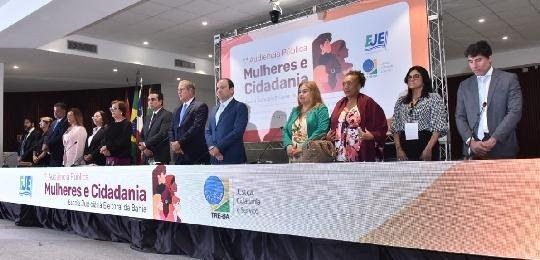 Pra todos verem: treze pessoas enfileiradas lado a lado, de pé, atrás de uma mesa de eventos.