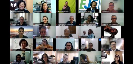 Pra todos verem: Captura de tela de uma videoconferência com cerca de 30 participantes distribuídos em janelas retangulares. As pessoas aparecem sorrindo, com variados cenários ao fundo, incluindo ambientes domésticos e institucionais. Algumas janelas exibem nomes e órgãos de origem.