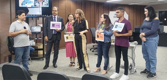 Pra todos verem: sete pessoas estão em pé em uma sala participando de uma atividade. Quatro delas seguram placas com números.
