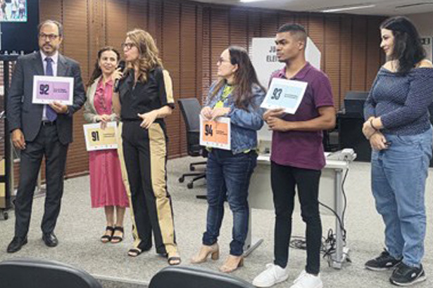 Pra todos verem: sete pessoas estão em pé em uma sala participando de uma atividade. Quatro delas seguram placas com números.
