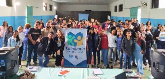 Pra todos verem: imagem com grupo de estudantes e servidores da EJE/BA sorriem em foto com placa do projeto ao centro.
