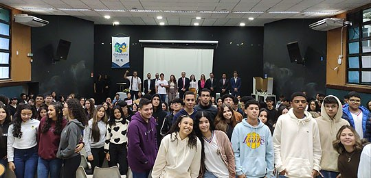 Pra todos verem: estudantes de ensino médio posam em auditório no lançamento de projeto, com telão e bandeira do projeto