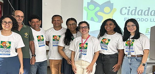 Cinco mulheres e três homens posa sorridente em frente a uma tela de projeção com o logotipo do projeto.